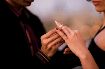 un homme vient de faire sa demande en mariage à sa fiancée à la période de noël. on voit l'homme passer la bague au doigt de sa femme. l'homme porte une veste noire avec une chemise avec des détails rouges. la femme porte un robe noire. la baque est argentée