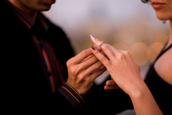 un homme vient de faire sa demande en mariage à sa fiancée à la période de noël. on voit l'homme passer la bague au doigt de sa femme. l'homme porte une veste noire avec une chemise avec des détails rouges. la femme porte un robe noire. la baque est argentée