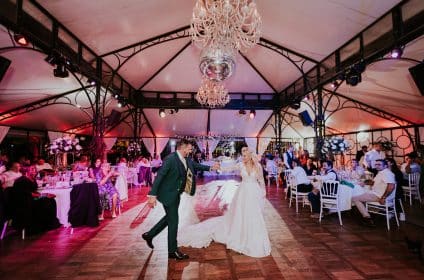 Deux mariés qui dansent en se tenant la main, dans une salle joliment décorée avec une lumière rosée. la mariée a une robe blanche et le marié un costume vert sapin