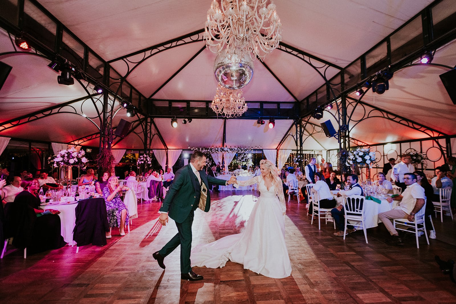 Deux mariés qui dansent en se tenant la main, dans une salle joliment décorée avec une lumière rosée. la mariée a une robe blanche et le marié un costume vert sapin